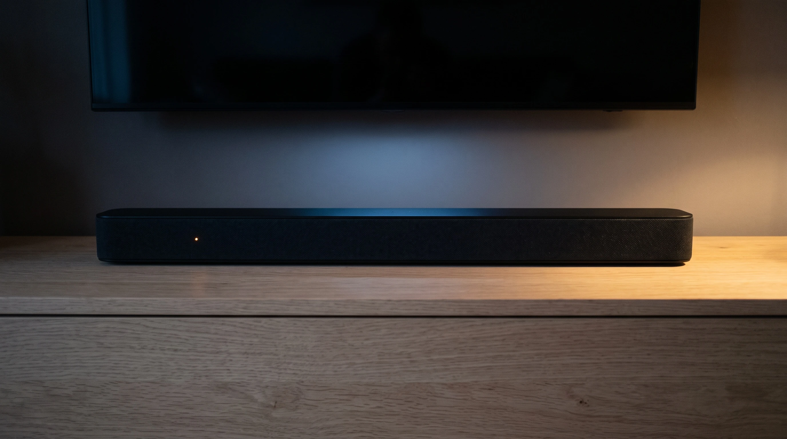 Närbild på en soundbar på en tv-bänk i ljus ek under en väggmonterad TV i ett mörkt rum